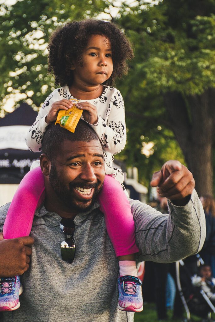 A joyful African American father with curly-haired daughter on shoulders, outdoors discovering Assentials. We are committed to educate, empower and elevate those that desire clarity, structure or simply want to preserve and secure what they have built - No matter where you are in your journey.