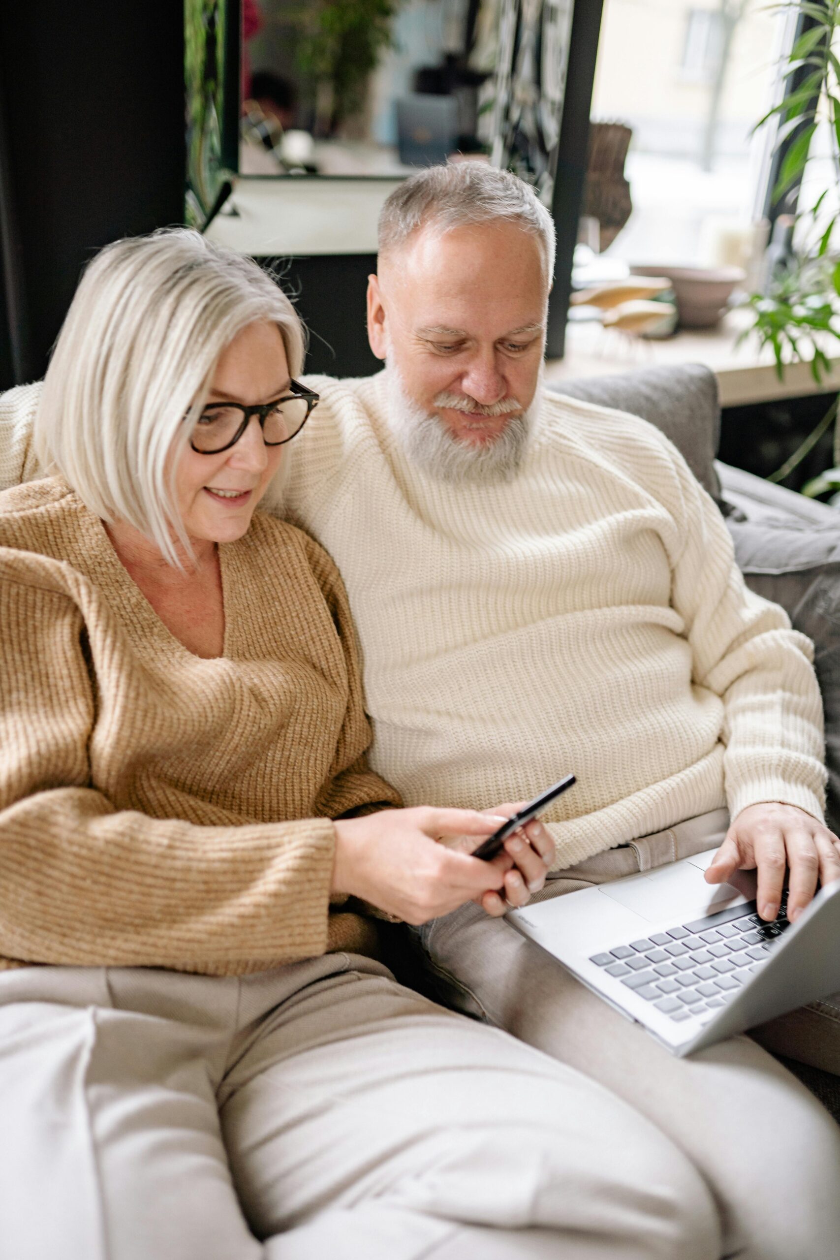 Senior couple using smartphone and laptop together on a cozy sofa indoors, depicting modern technology lifestyle.